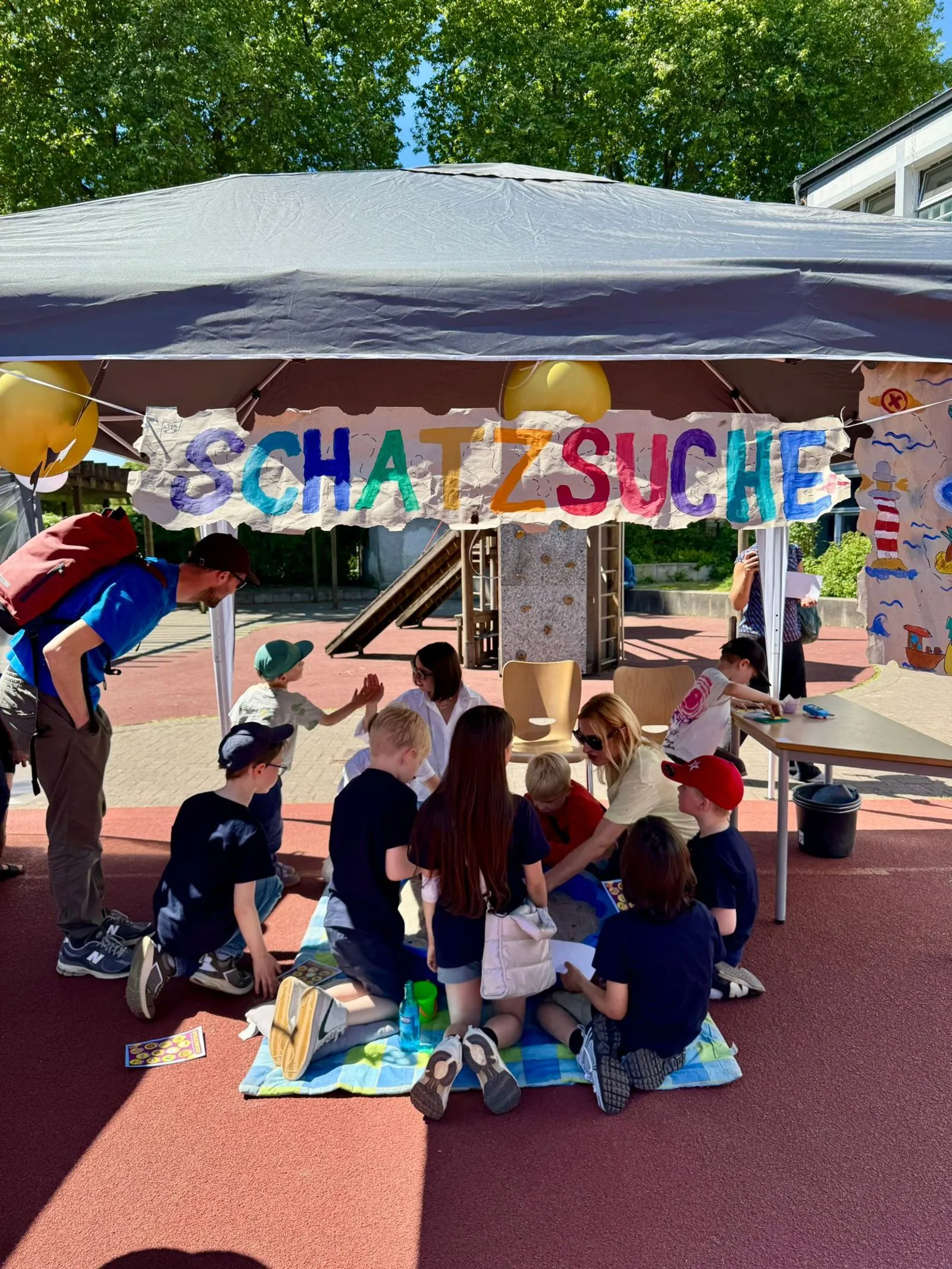 Pavillon mit buntem "SCHATZSUCHE"-Banner, mehrere Kinder sitzen auf Decke und basteln, Spielplatz im Hintergrund