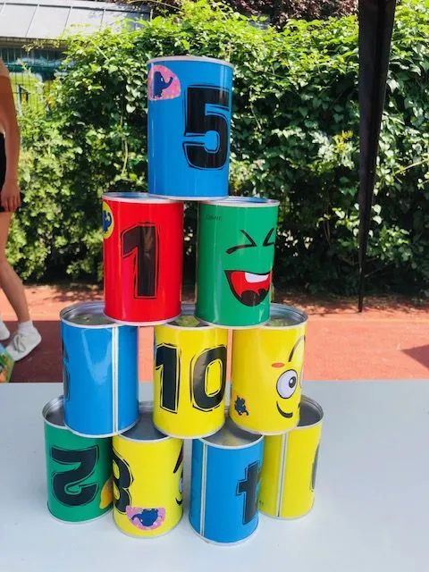 Pyramide aus bunten Dosen mit Zahlen und Comic-Gesichtern auf Tisch vor grünem Gebüsch