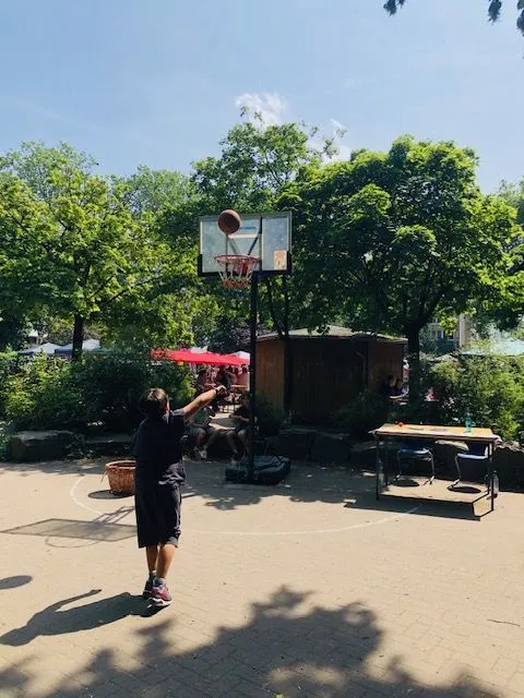 Junge wirft Basketball auf Freiluftkorb in sonnigem Park mit Bäumen und Tischen