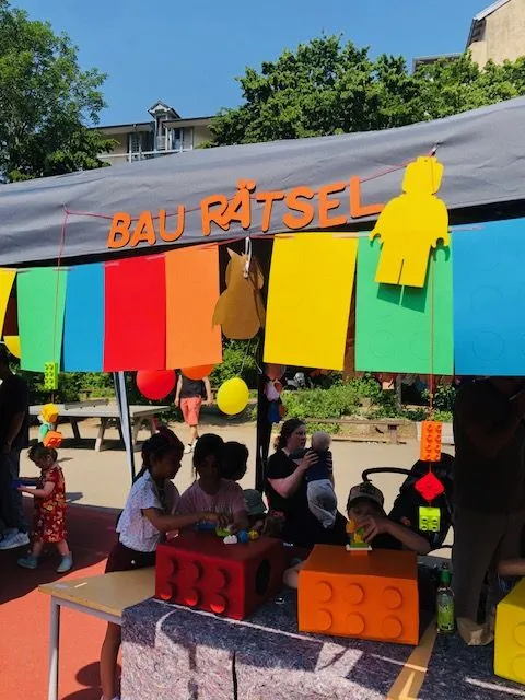 Buntes Standzelt mit Schrift "BAU RÄTSEL", Kinder spielen an großen LEGO‑ähnlichen Bausteinen