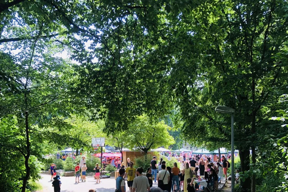 Menschen bei Sommerfest im schattigen Park mit Marktständen, spielenden Kindern, Kinderwagen und Bäumen