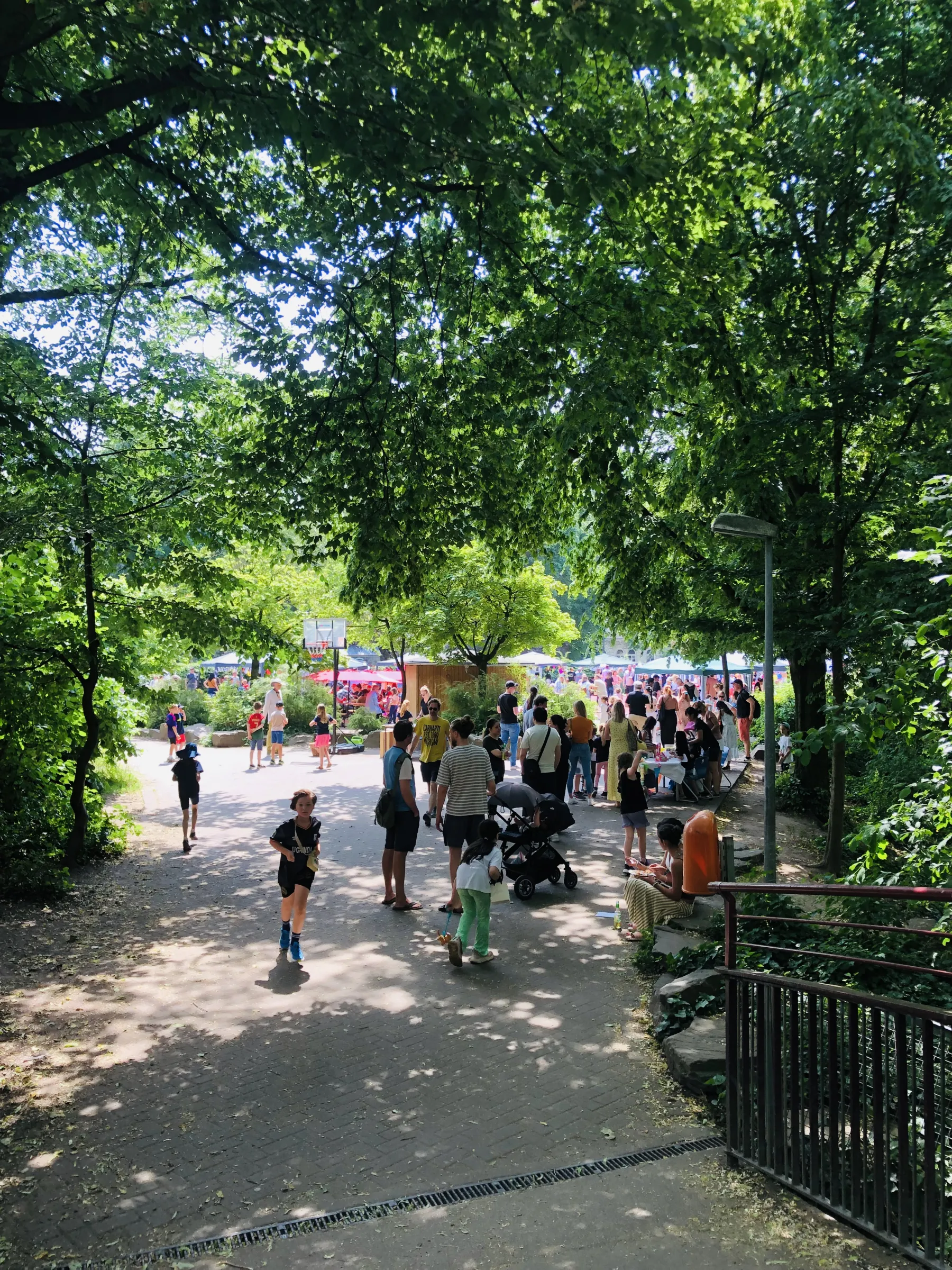 Menschen bei Sommerfest im schattigen Park mit Marktständen, spielenden Kindern, Kinderwagen und Bäumen