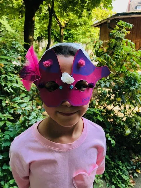 Mädchen mit pink-violetter Bastelmaske mit Federn und Perlen vor grünen Gartensträuchern