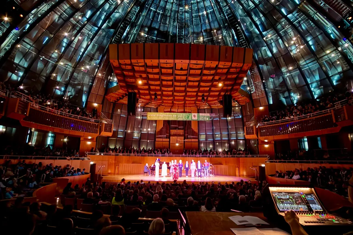 Große Konzerthalle mit gläserner Kuppel, beleuchteter Bühne mit Ensemble und vollbesetztem Publikum