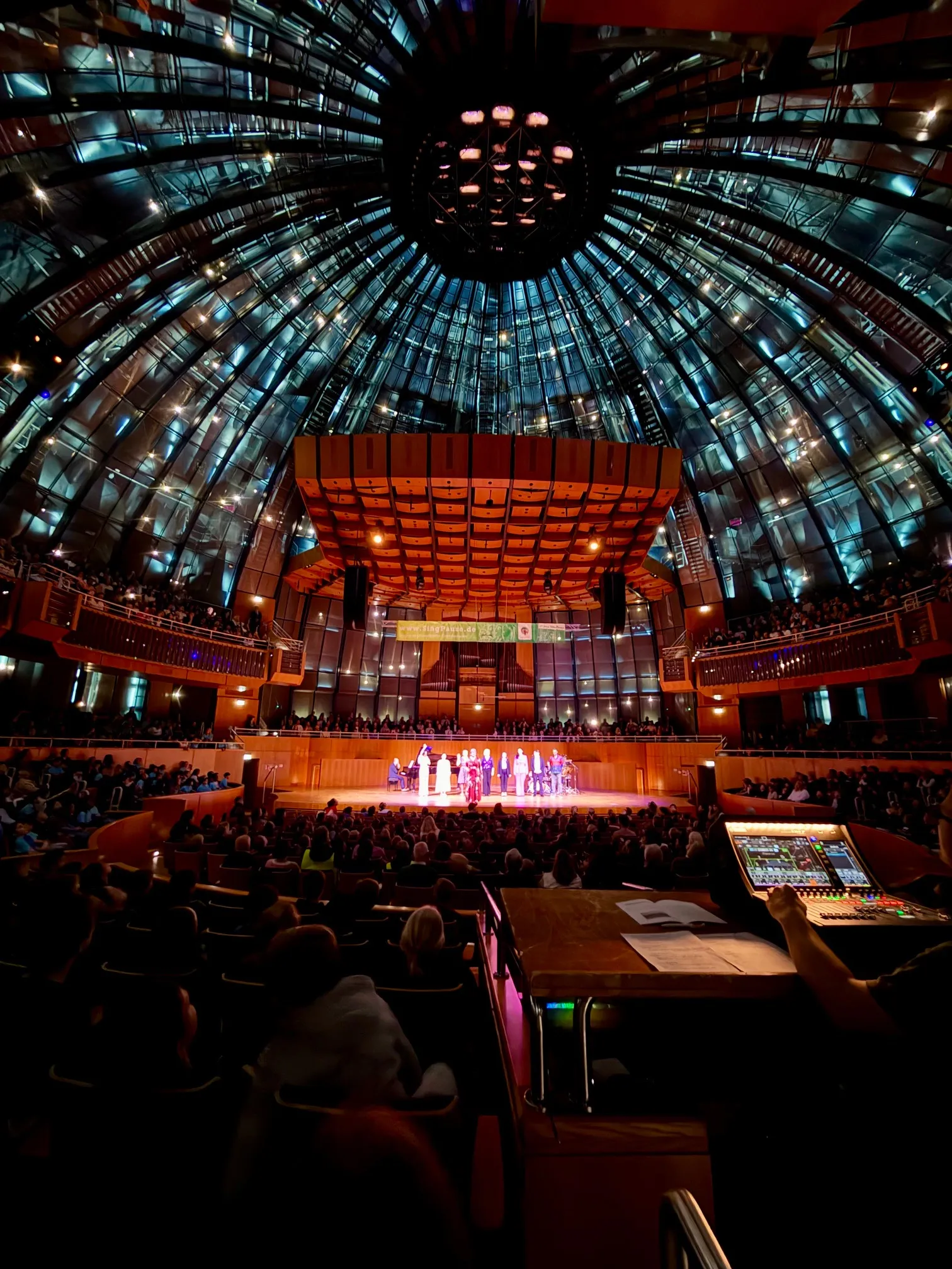 Große Konzerthalle mit gläserner Kuppel, beleuchteter Bühne mit Ensemble und vollbesetztem Publikum
