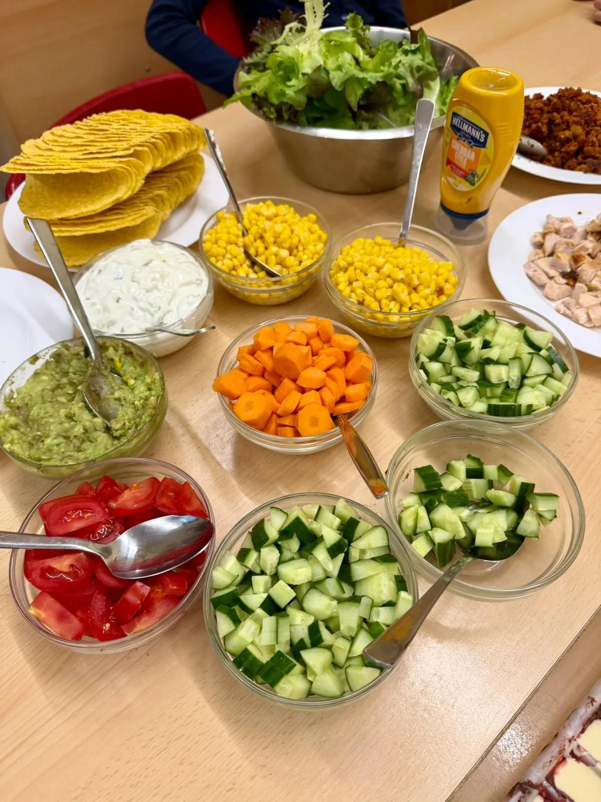 Tisch mit Tortillawraps, Schüssel grüner Salat und mehrere Schälchen mit Mais, Karotten, Gurke, Tomate, Guacamole und Dips.