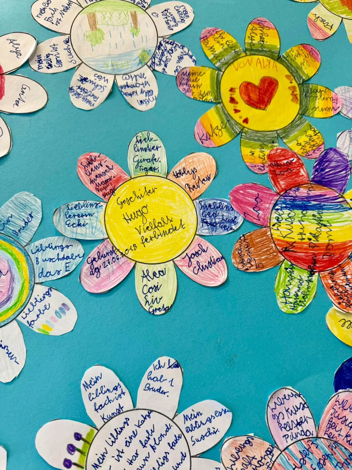 Bunte, handgemalte Papierblumen mit beschrifteten Blütenblättern auf türkisfarbenem Hintergrund.