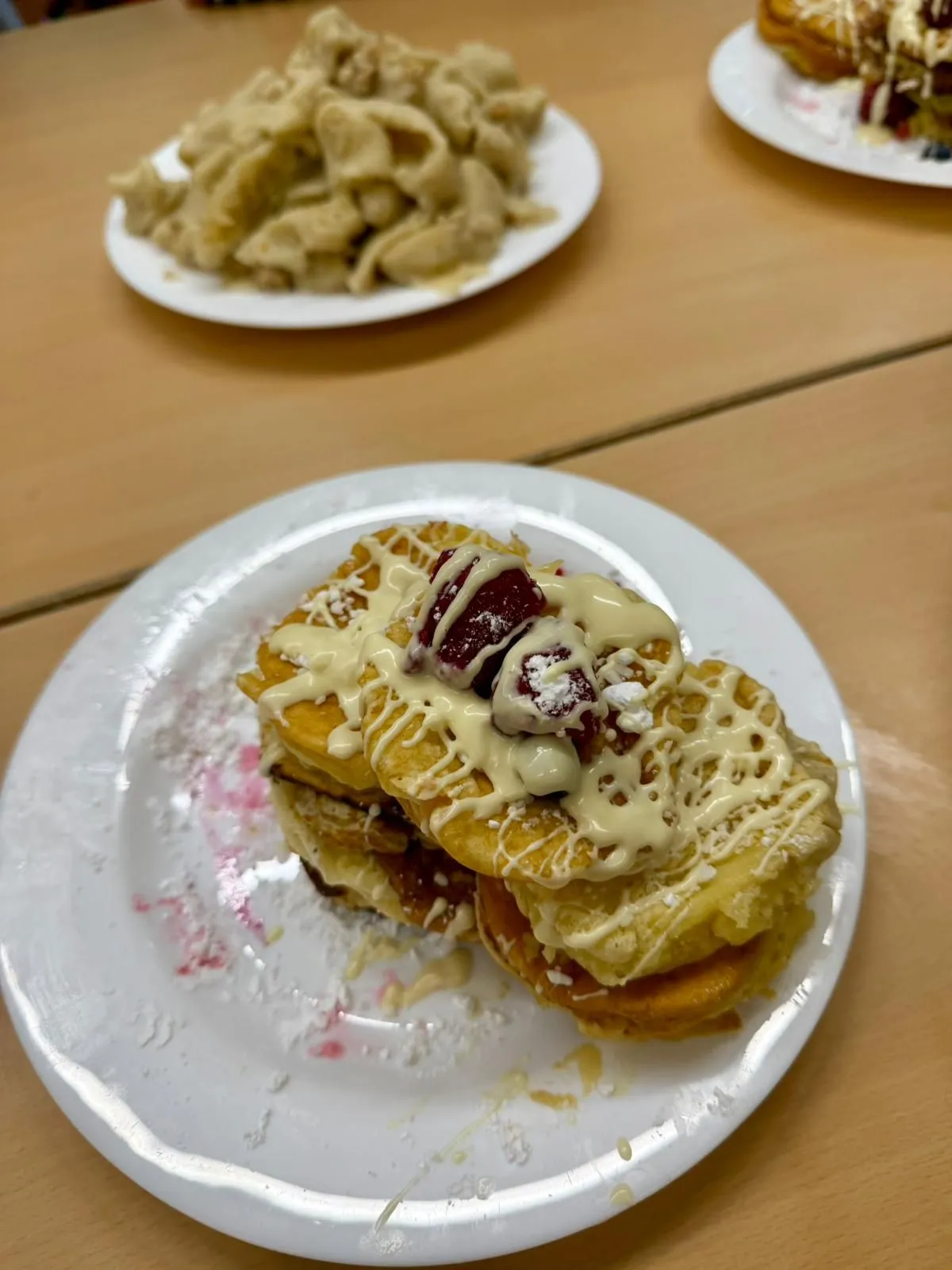 Gestapelte Pfannkuchen mit weißer Schokoladensauce und Himbeeren auf weißem Teller, im Hintergrund weiterer Teller