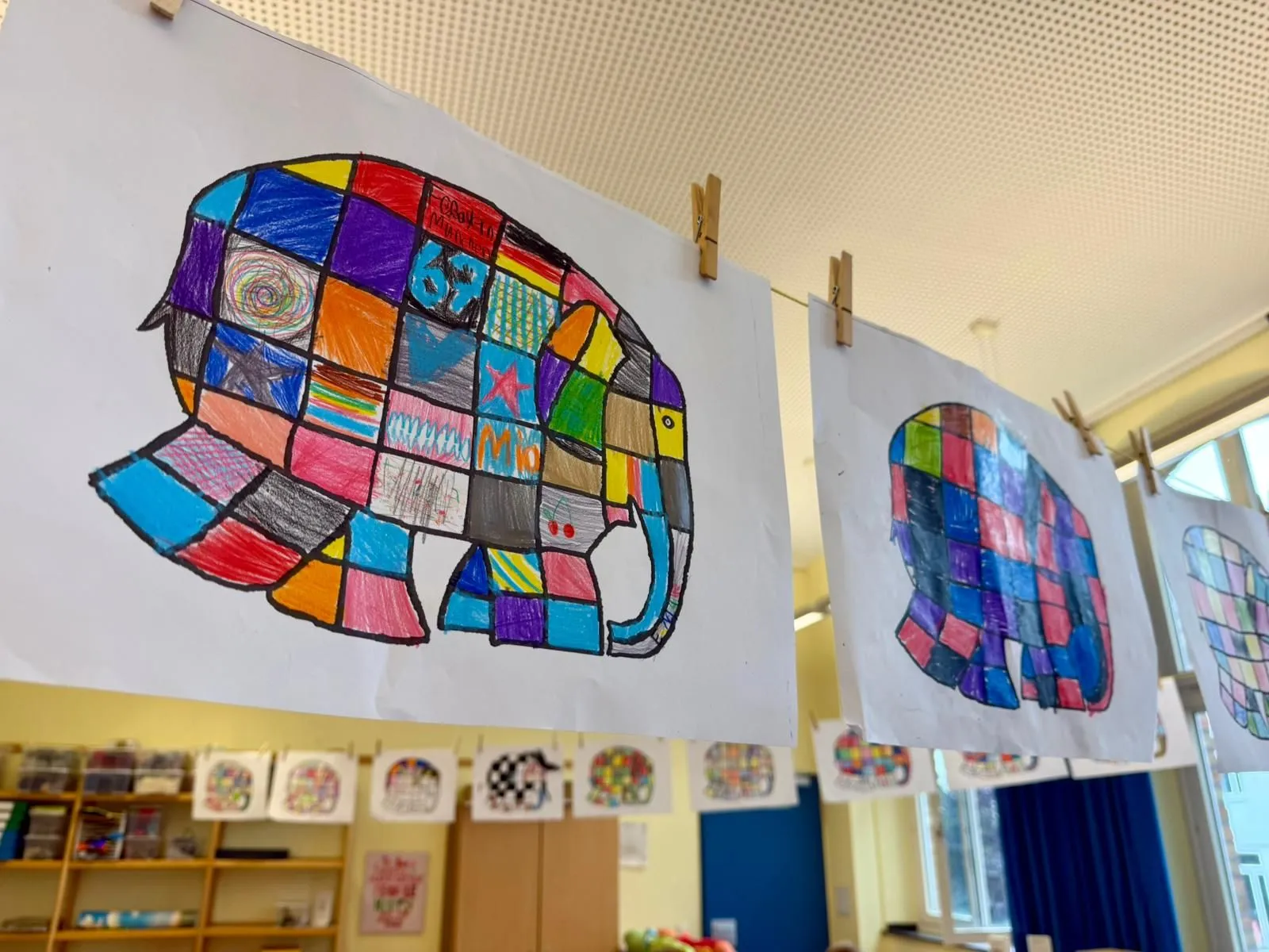 Bunte kariert ausgemalte Elefanten auf Papier an einer Schnur mit Wäscheklammern im Klassenraum