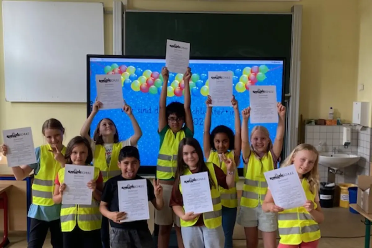 Kinder in gelben Warnwesten halten Urkunden vor Tafel mit buntem Luftballon-Hintergrund im Klassenraum