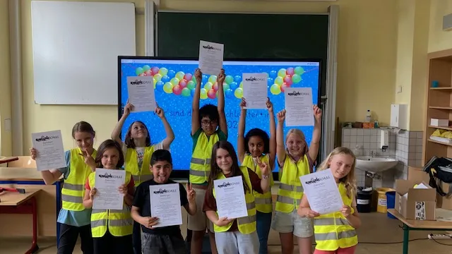 Kinder in gelben Warnwesten halten Urkunden vor Tafel mit buntem Luftballon-Hintergrund im Klassenraum