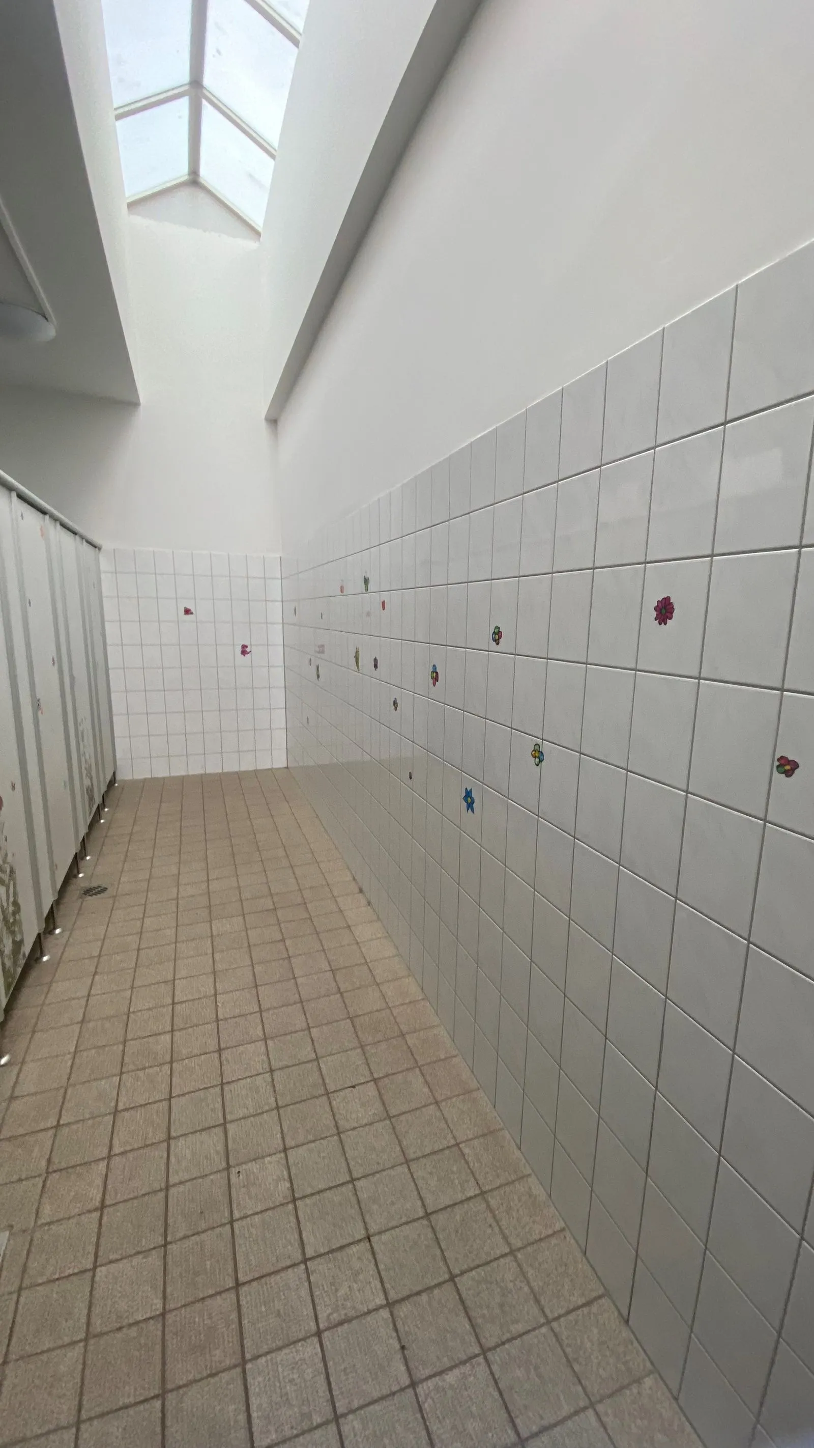 Flur mit weißen Wandfliesen und kleinen bunten Blumenstickern, Toilettenkabinen links und Oberlicht oberhalb