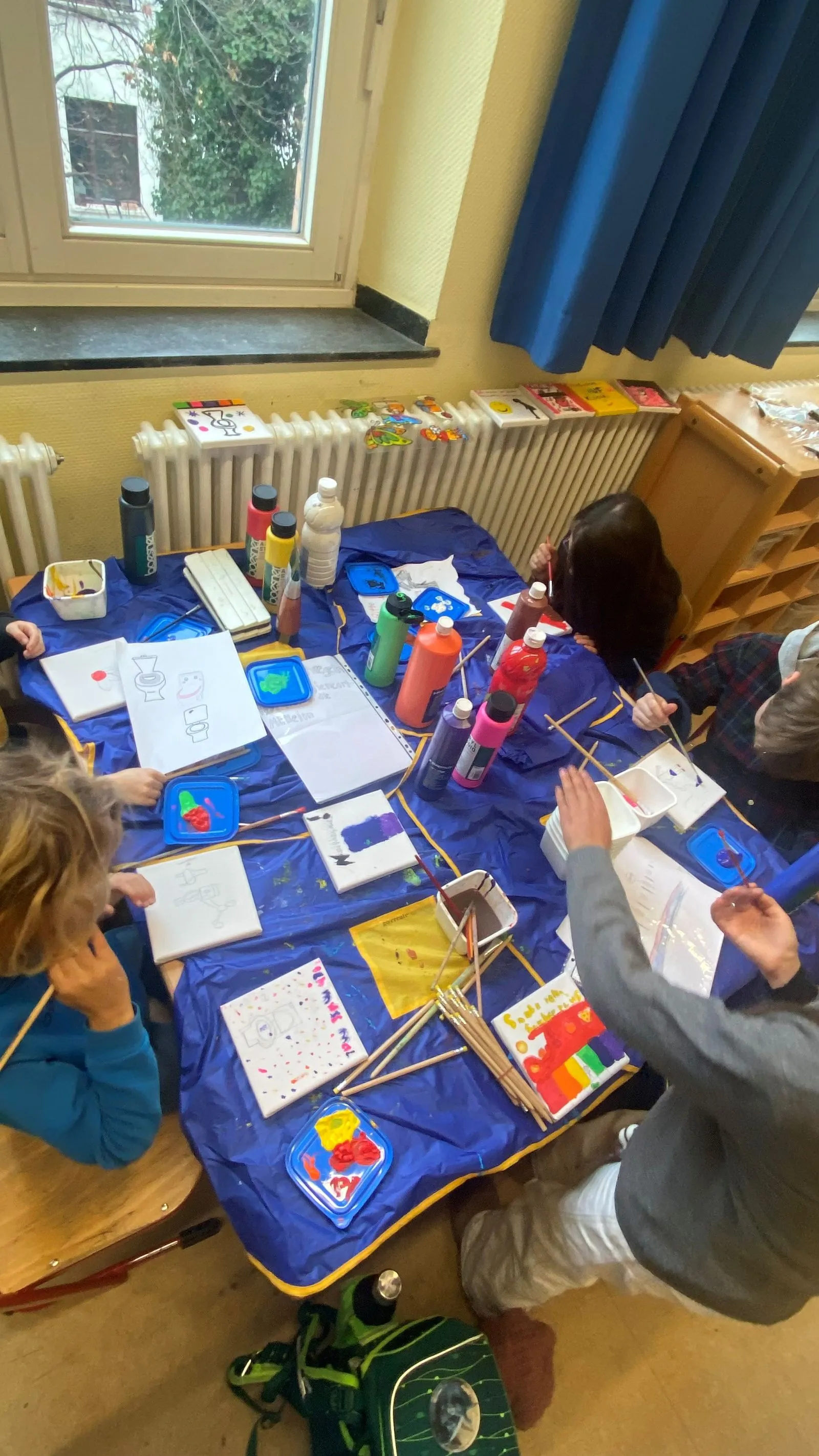 Mehrere Kinder malen an einem Tisch mit Leinwänden, Pinseln, bunten Farbflaschen und blauer Tischdecke neben Fenster