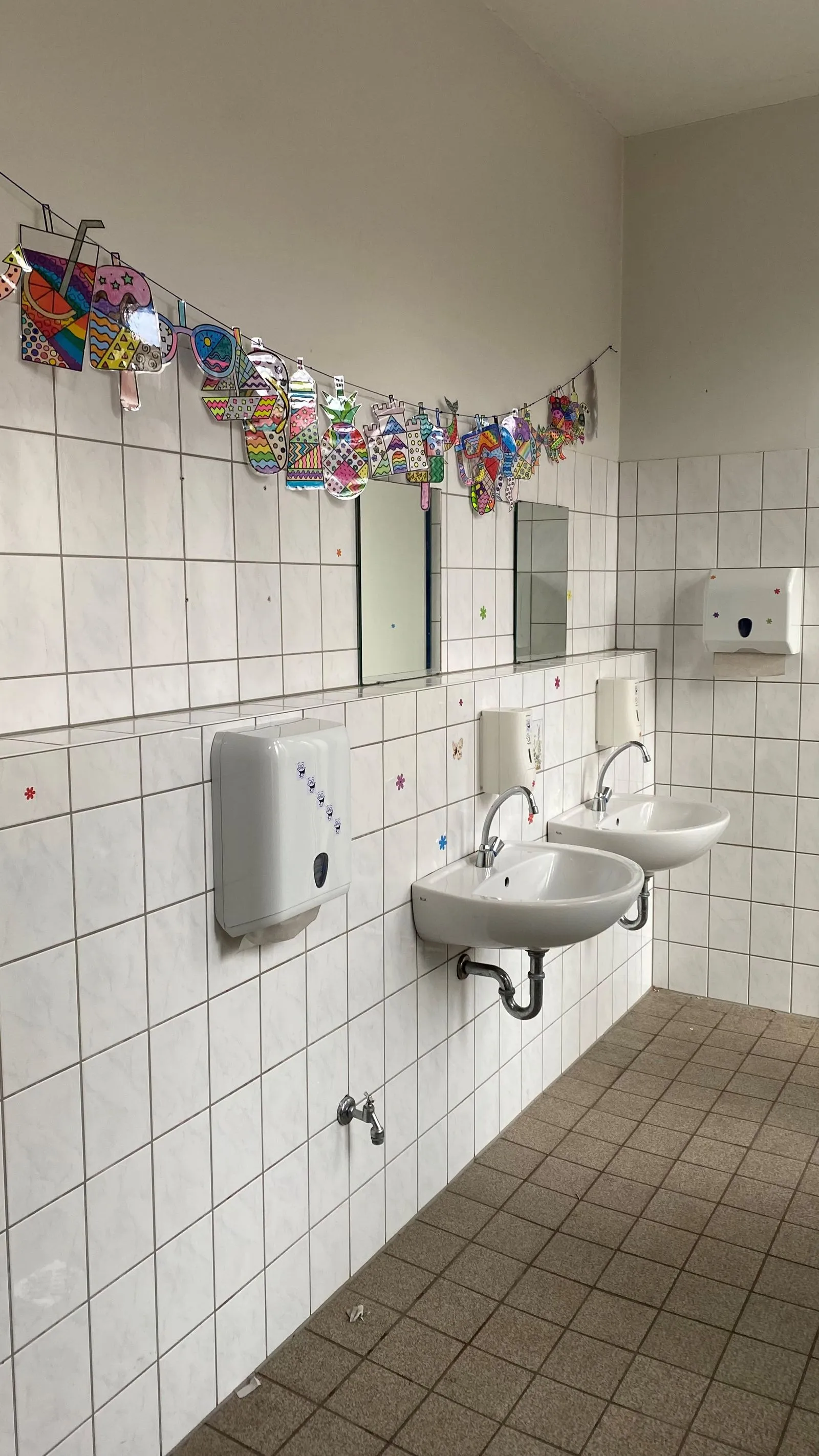 Weiße Kachelwand mit zwei Waschbecken, Seifenspendern, Spiegeln und bunten Kinderzeichnungen an einer Schnur