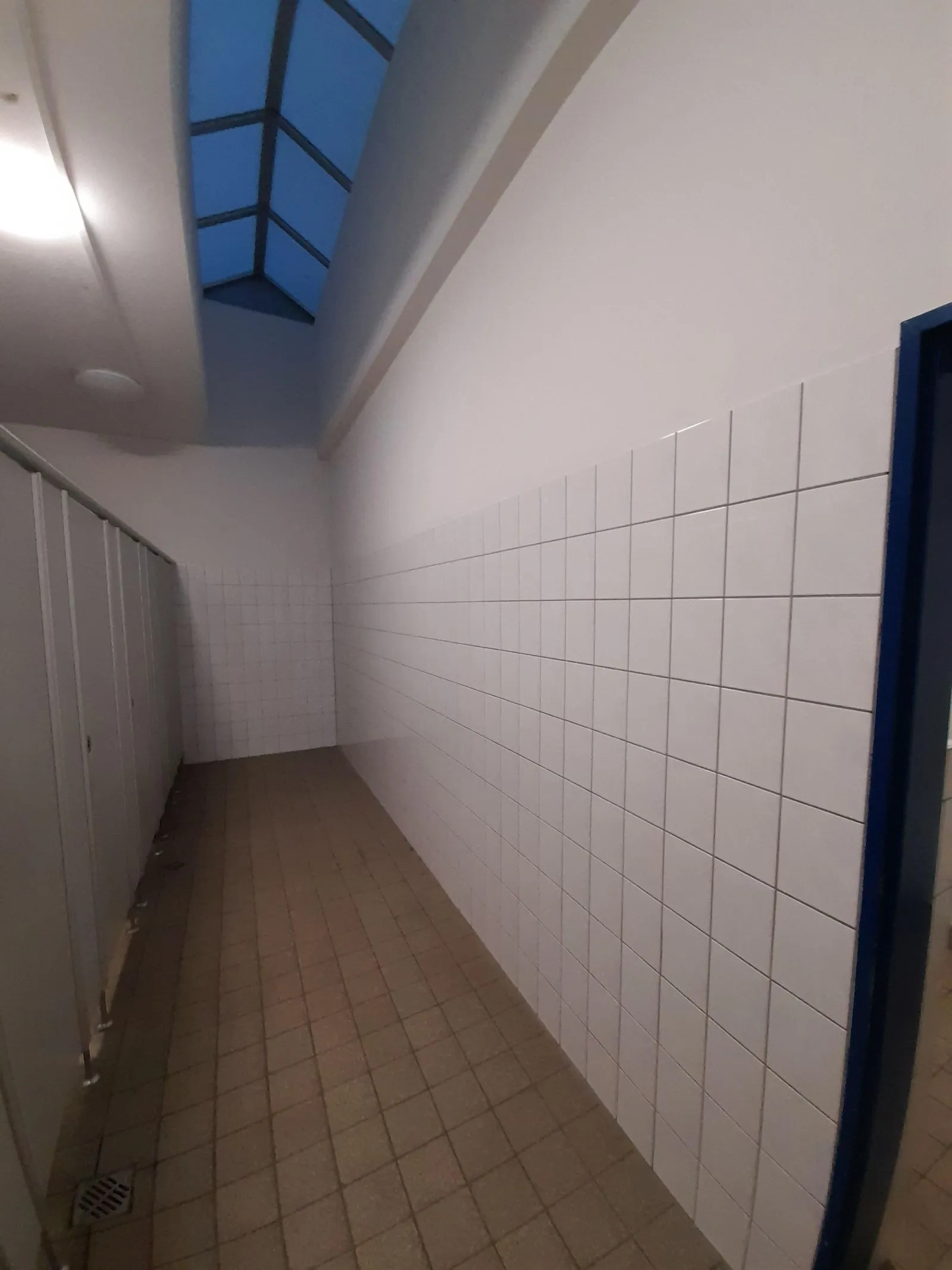 Schmaler Flur mit weißen Wandfliesen und beigen Bodenfliesen, links Toilettenkabinen, oben großes Dachfenster