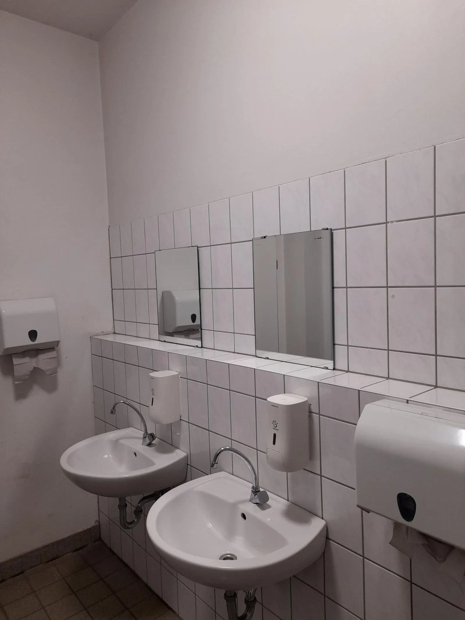 Öffentliche Toilettenwand mit zwei weißen Waschbecken, Seifenspendern, zwei Spiegeln und Papierhandtuchspendern
