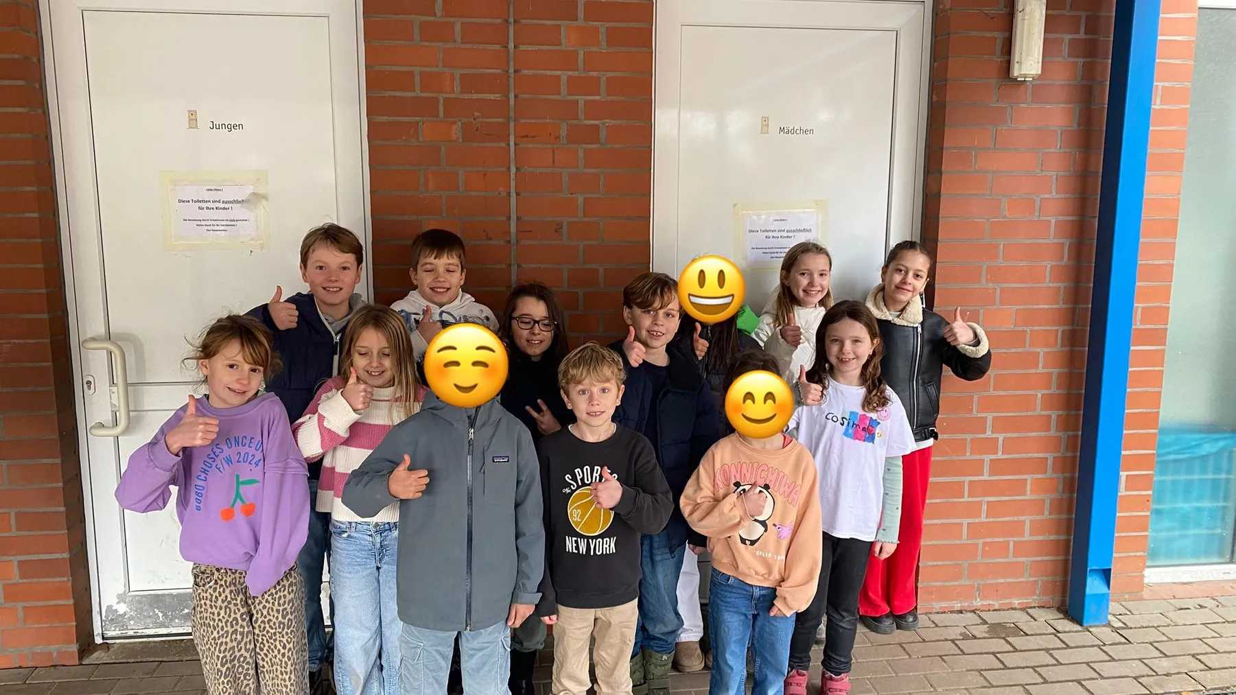 Gruppe Kinder vor roter Backsteinwand und zwei Türen mit Schildern „Jungen“ und „Mädchen“, einige zeigen Daumen hoch
