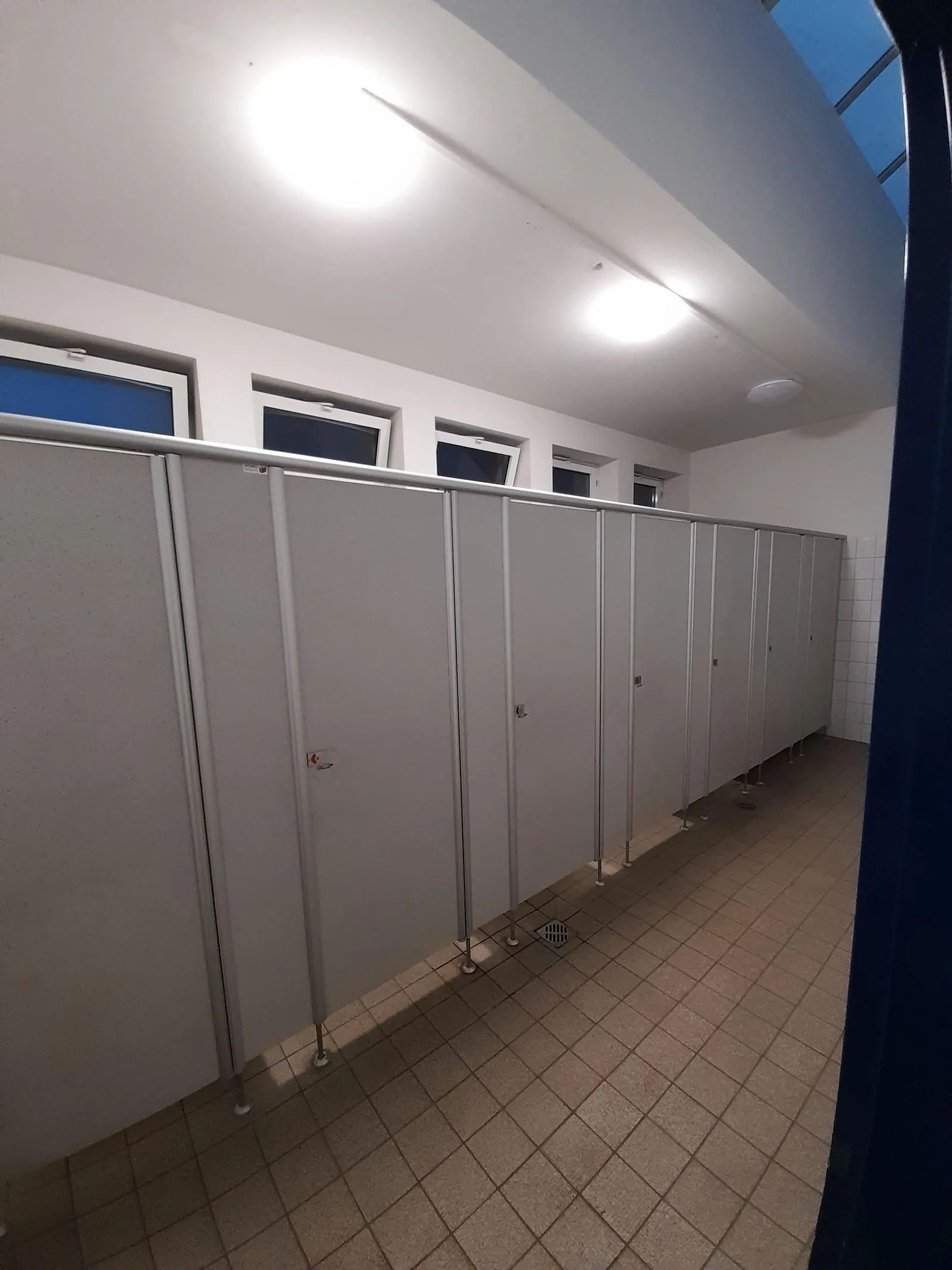 Reihe geschlossener grauer WC-Kabinen in hell beleuchtetem Sanitärraum mit Kachelboden und oberer Fensterreihe