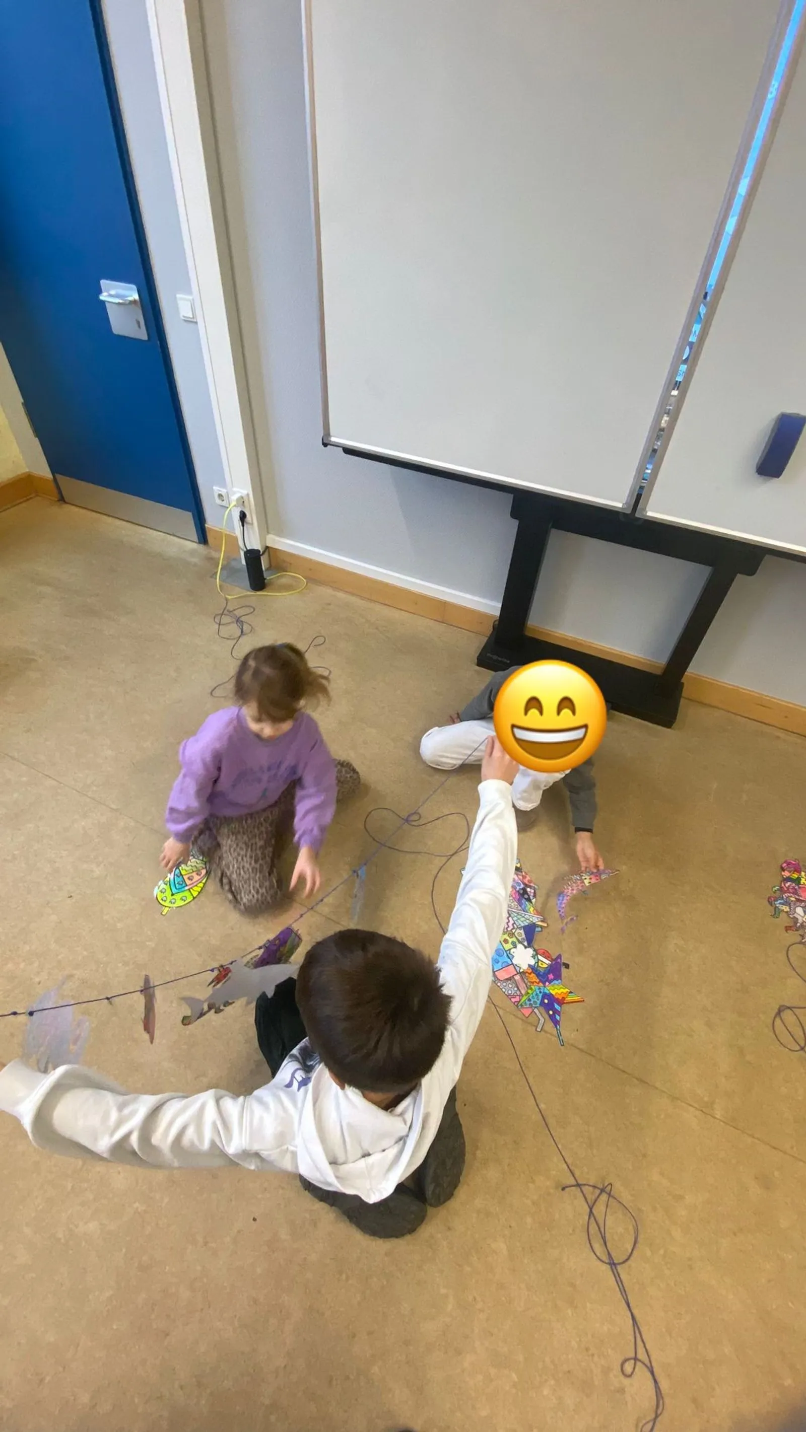 Drei Kinder sitzen auf dem Boden und ordnen bunte, gemusterte Papierfische an einer Schnur vor einer weißen Tafel