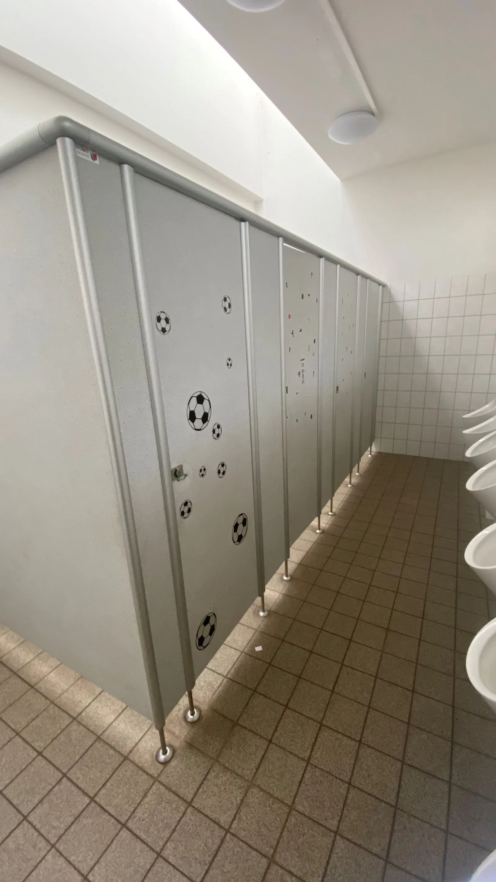 Reihe grauer WC-Kabinen mit Fußballaufklebern, Kachelwand, braun-graue Bodenfliesen und sichtbare Urinale rechts