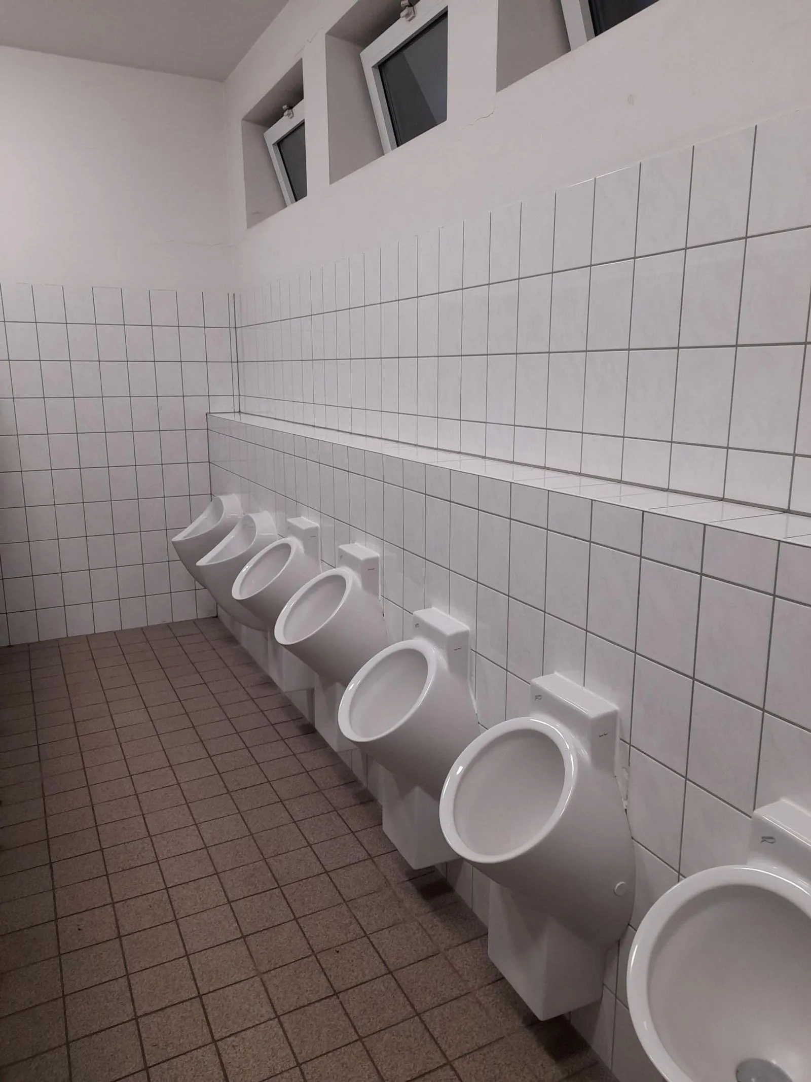 Reihe von sieben weißen Urinalen an weiß gefliester Wand, brauner gefliester Boden und drei hohe Kippfenster oben