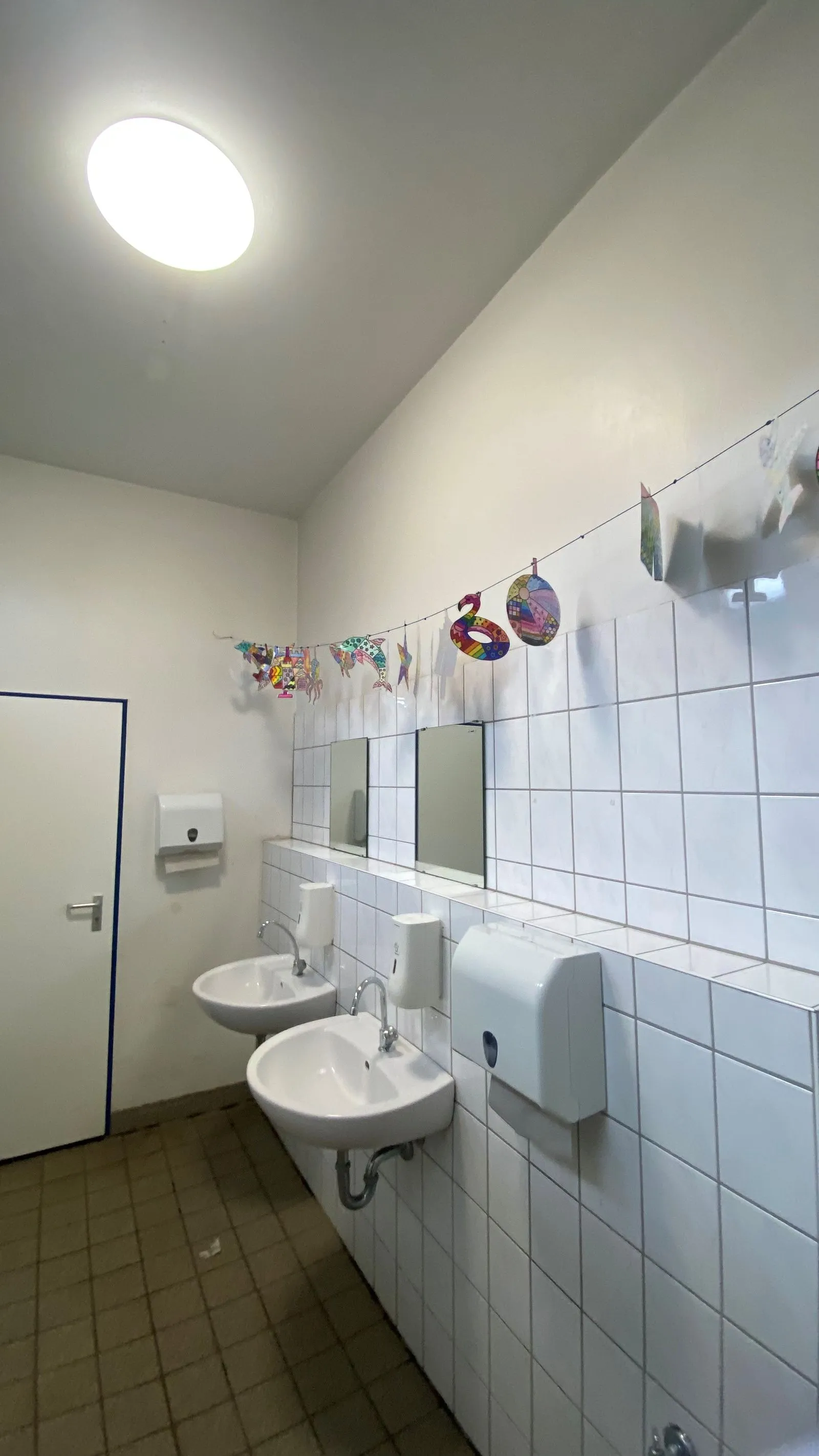 Zweifach-Waschbecken an gefliester Wand mit Spiegeln, Seifen- und Papierspendern und aufgehängten bunten Papierdekorationen.