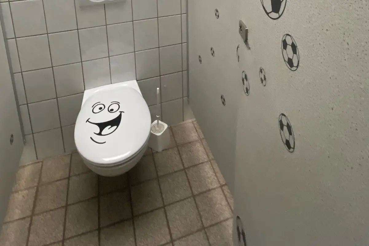 Toilette mit lächelndem Sitzdeckel-Aufkleber, geflieste Wand, Bodenfliesen und Fußball-Aufkleber auf Trennwand