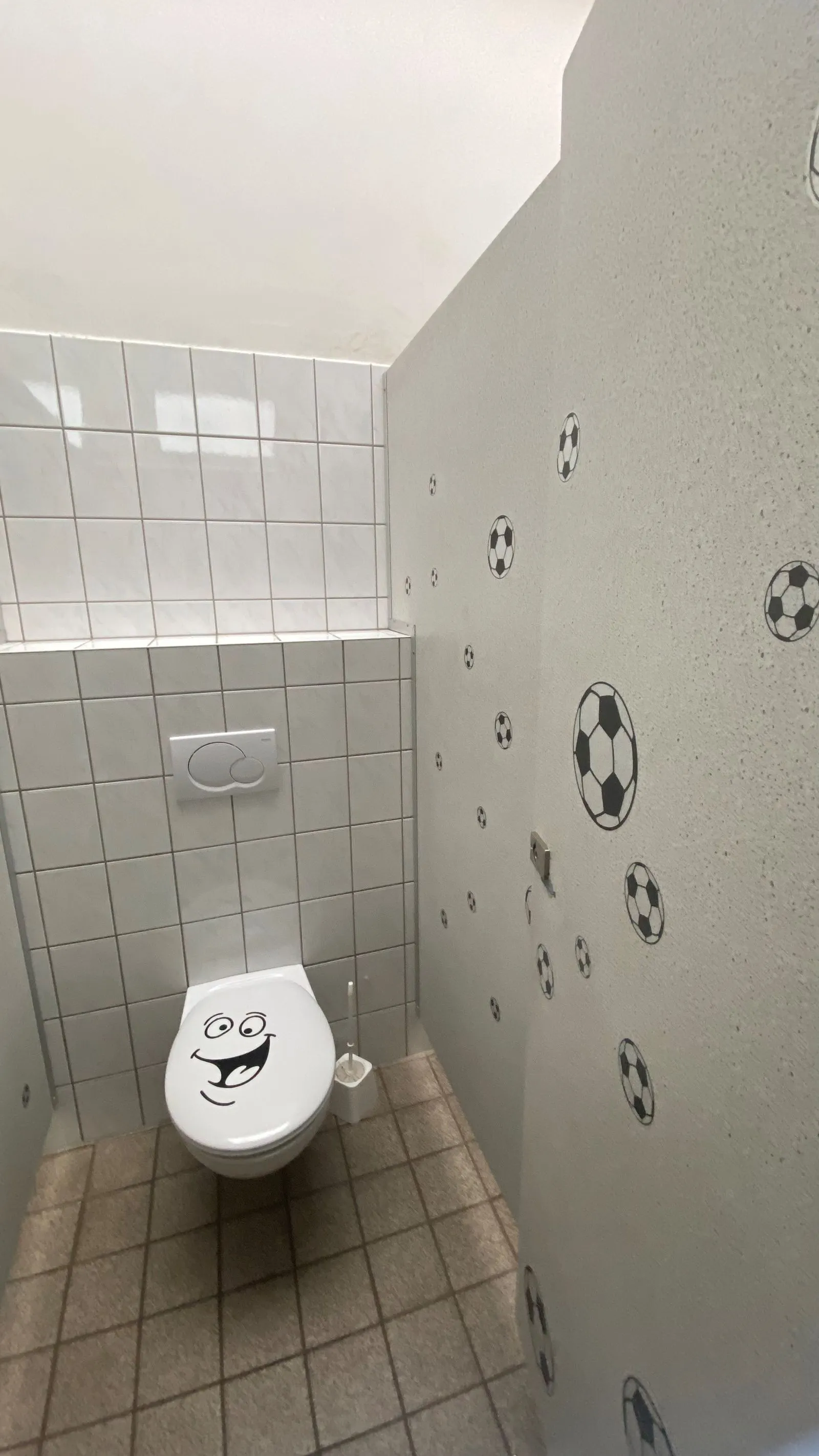 Toilette mit lächelndem Sitzdeckel-Aufkleber, geflieste Wand, Bodenfliesen und Fußball-Aufkleber auf Trennwand