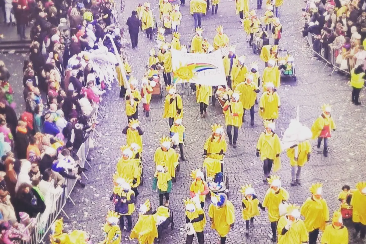 Karnevalsumzug aus Vogelperspektive: Menschen in gelben Umhängen mit Sonnenkopfschmuck, Regenbogenbanner, Zuschauer an Absperrungen
