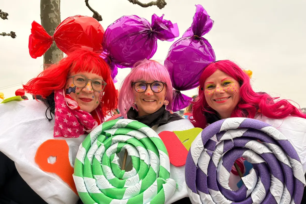 Drei kostümierte Personen mit bunten Perücken, Gesichtsfarbe, großen Lutscher-Requisiten und glänzenden Bonbon‑Hüten