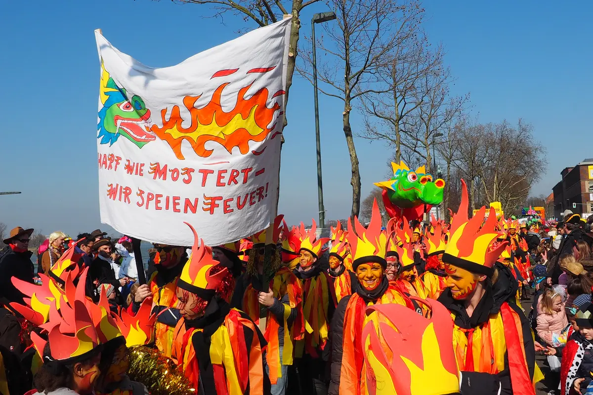 Karnevalsgruppe in feuerfarbenen Kostümen und Flammenhüten, große Fahne mit feuerspuckendem Drachen