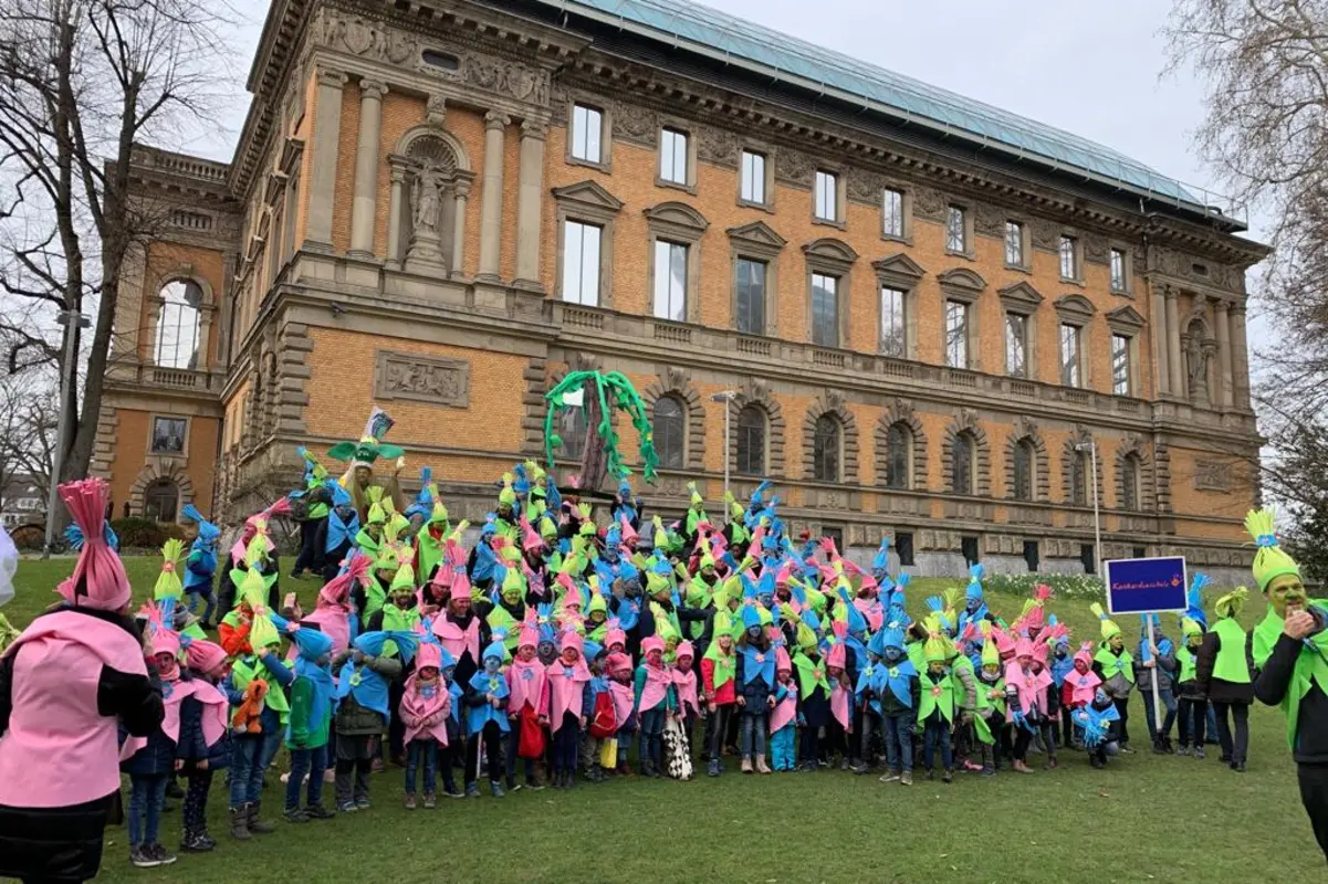 Große Gruppe verkleideter Kinder und Erwachsener in neonpinken, -grünen und blauen Spitzhüten vor historischem Backsteingebäude