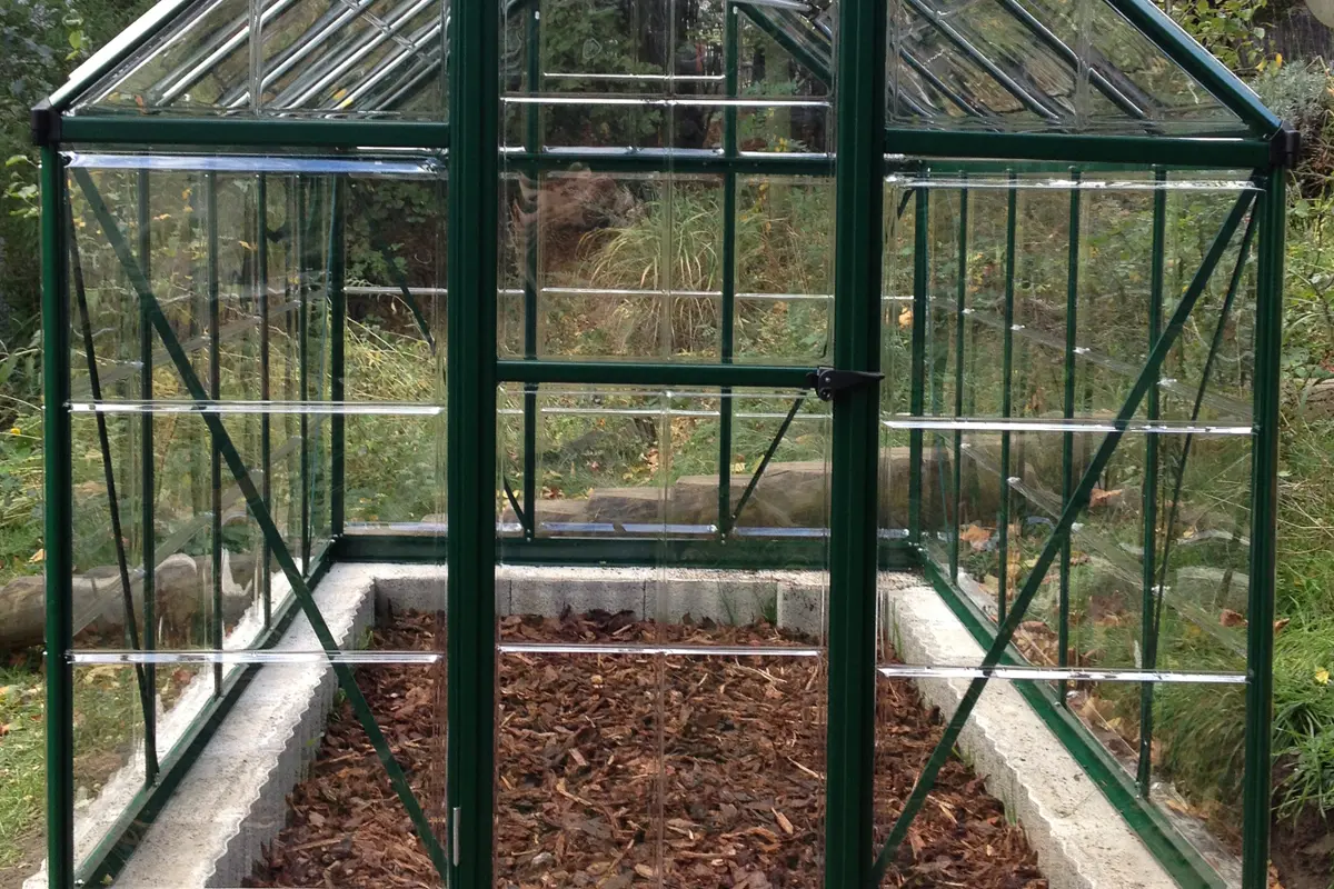 Grünes Glasgewächshaus auf Betonsockel, innen mit Laubmulch bedeckt, umgeben von Bäumen und Sträuchern