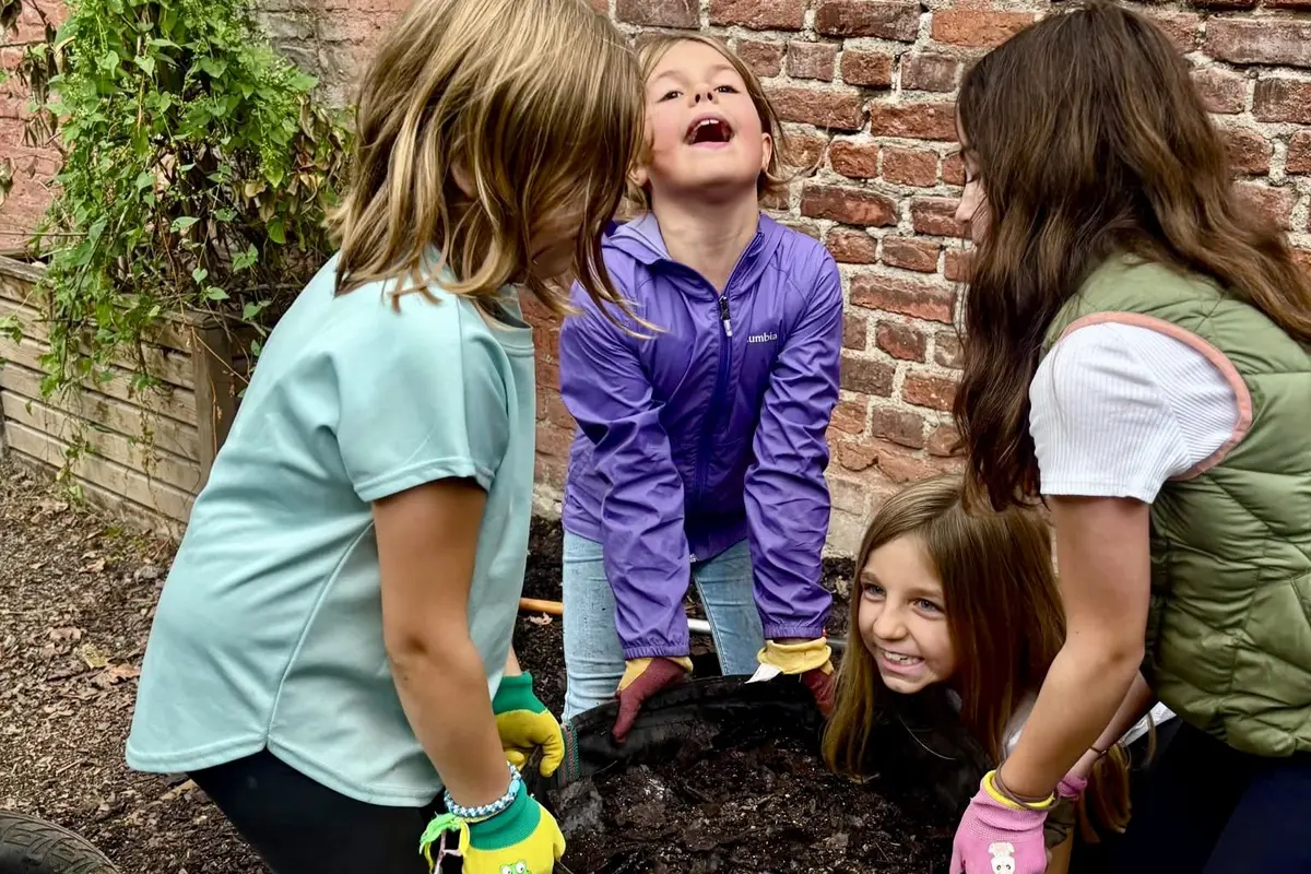 Vier Kinder mit bunten Gartenhandschuhen heben gemeinsam einen großen Pflanzkübel mit Erde vor einer Backsteinmauer