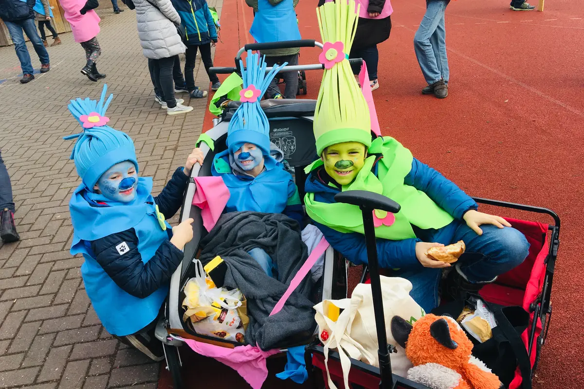Drei Kinder mit blauer und grüner Gesichtsbemalung und bunten Filzhüten in Bollerwagen