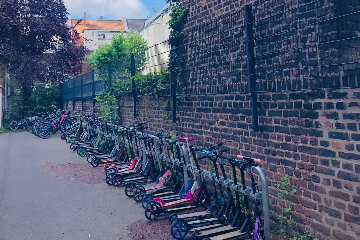 Viele geparkte E‑Scooter in einer Reihe neben hoher Backsteinmauer und Drahtzaun, Bäume und Häuser im Hintergrund
