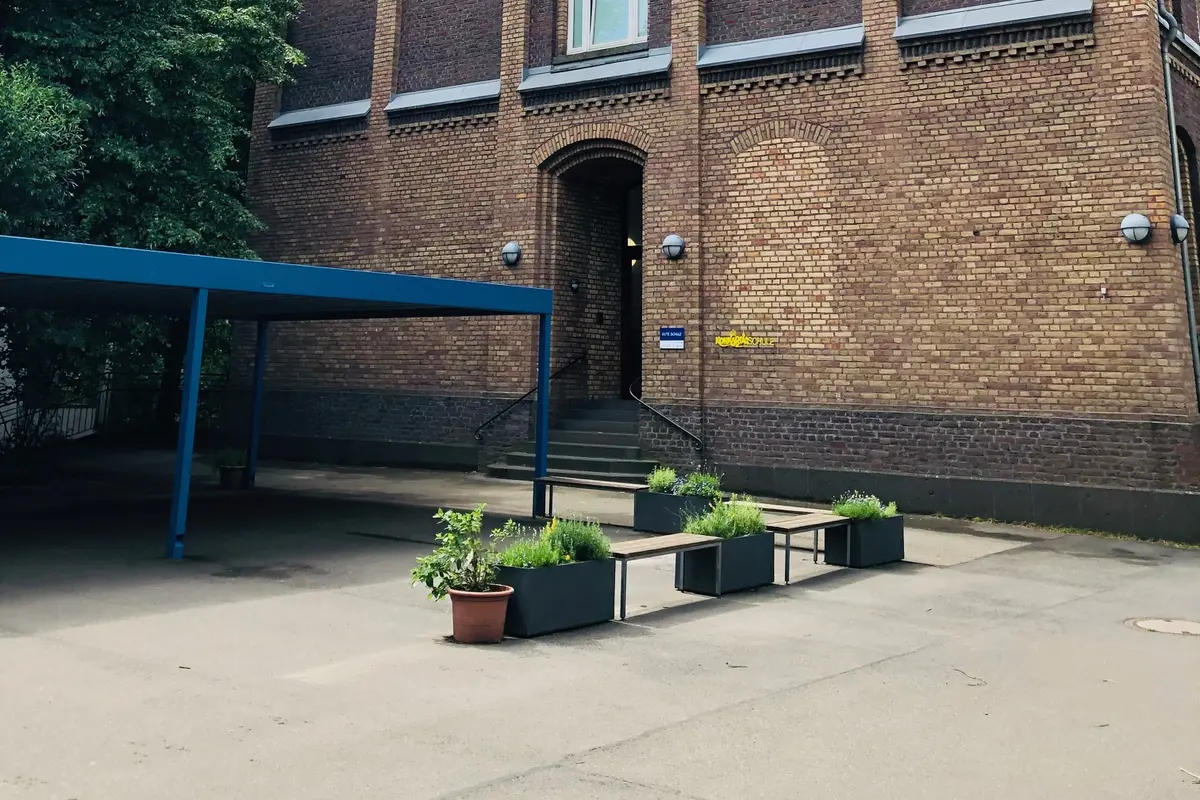 Backsteingebäude mit drei hohen Fensterachsen, Torbogen-Eingang, blauem Vordach und Pflanzkästen mit Bänken im Hof
