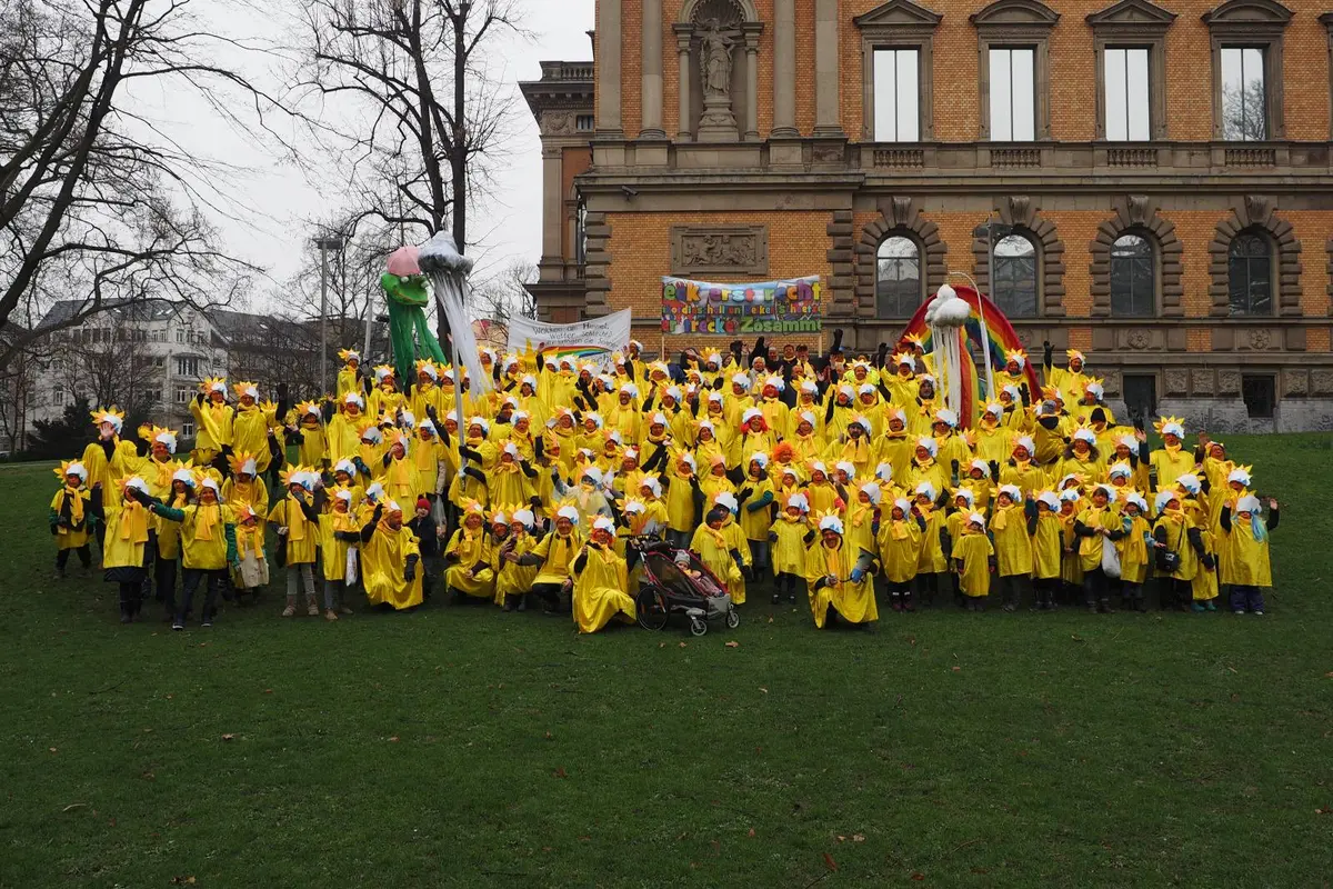 Große Gruppe in gelben Regenponchos mit weißen Blumenhüten vor historischem Gebäude, bunte Banner und Requisiten
