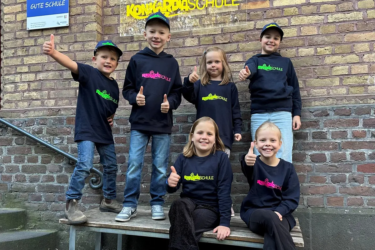 Sechs Kinder in schwarzen Pullovern und Baseballmützen vor Backsteinwand zeigen den Daumen hoch
