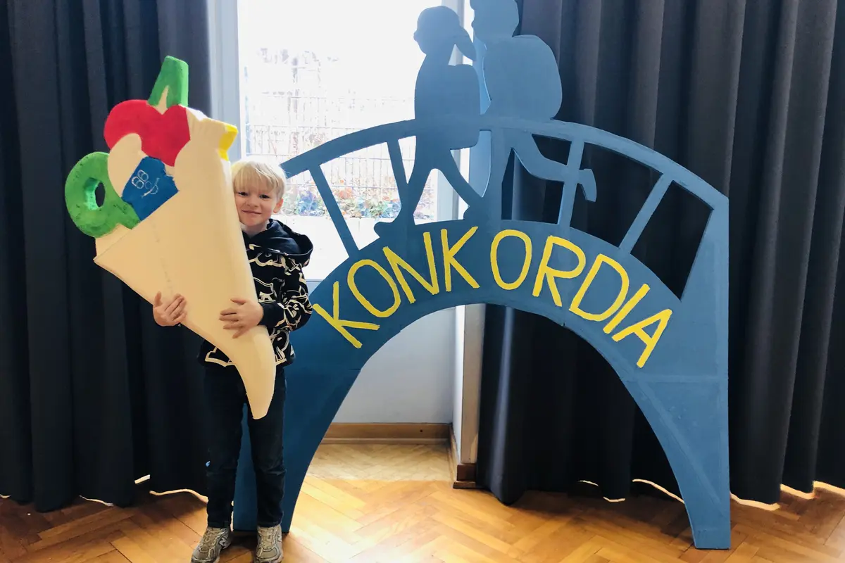Blonder Junge hält große bunte Schultüte vor blauem Deko-Bogen mit gelber Aufschrift "KONKORDIA"