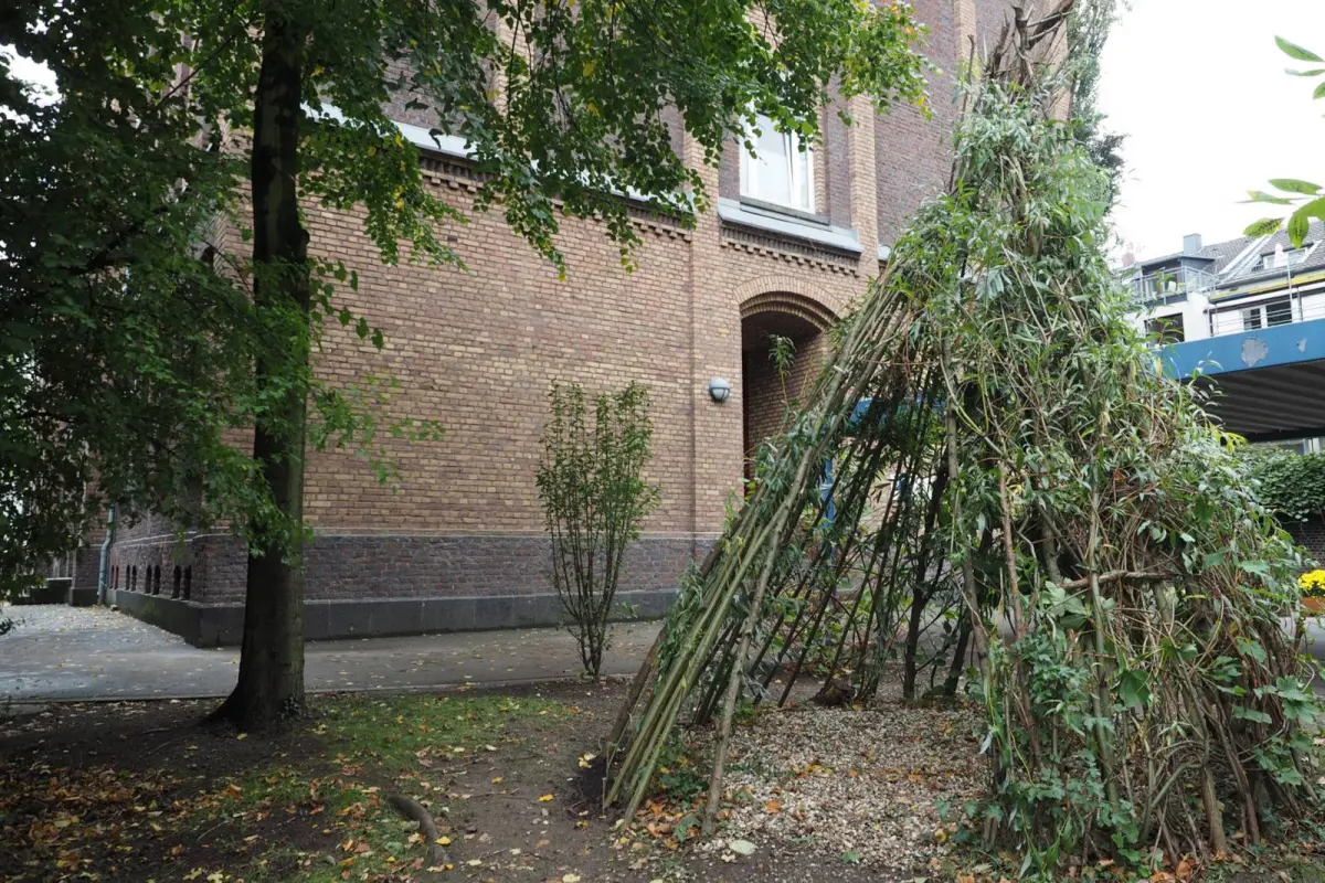 Weiden-Tipi aus Zweigen und Blättern neben einem Backsteingebäude und Bäumen