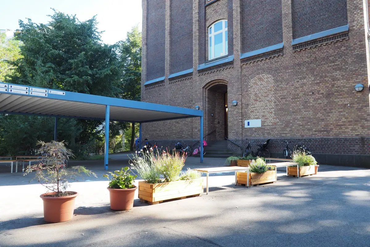 Hof vor rotem Backsteingebäude mit überdachtem Fahrradstand, Fahrrädern, Pflanzkübeln und Bänken.
