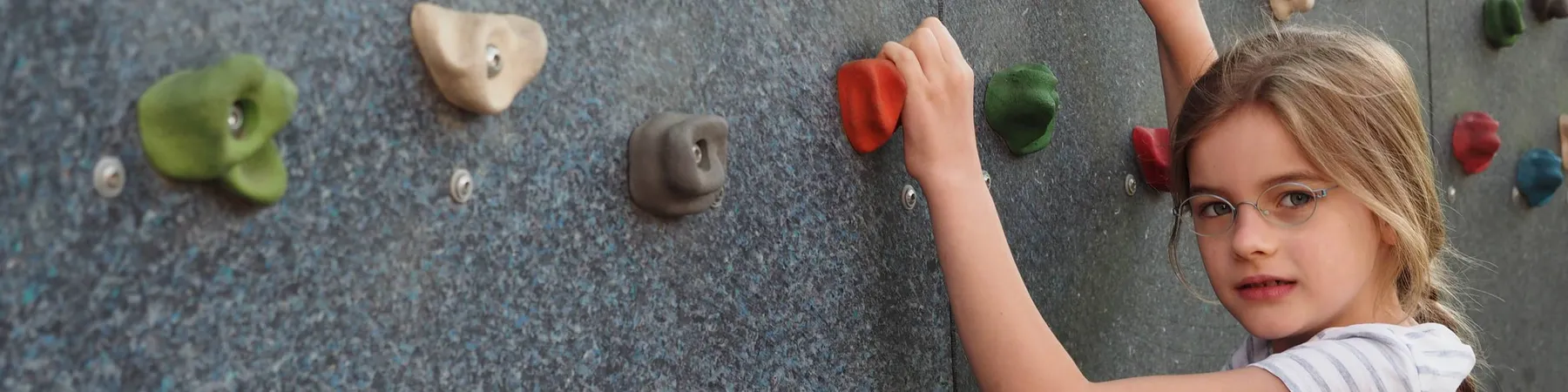 Mädchen mit Brille klettert an einer Kletterwand und greift rote und grüne Griffsteine