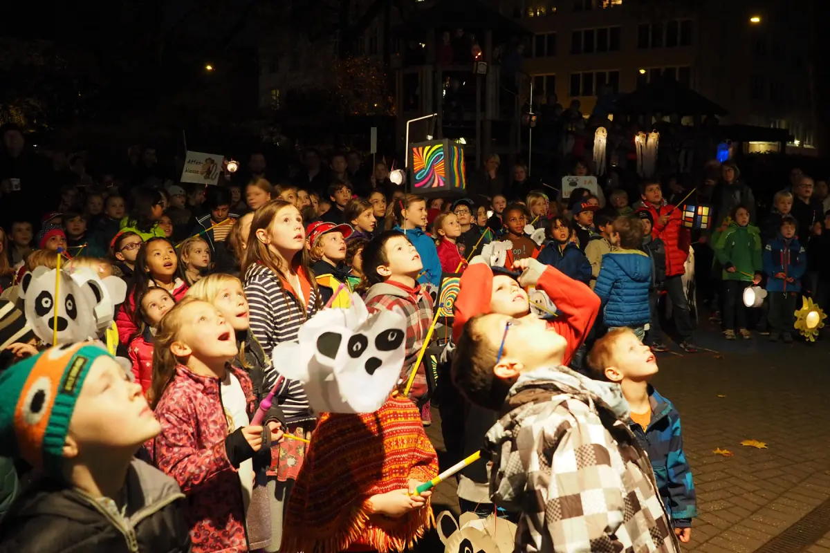Viele Kinder mit bunten Laternen blicken bei einer Nachtveranstaltung nach oben.