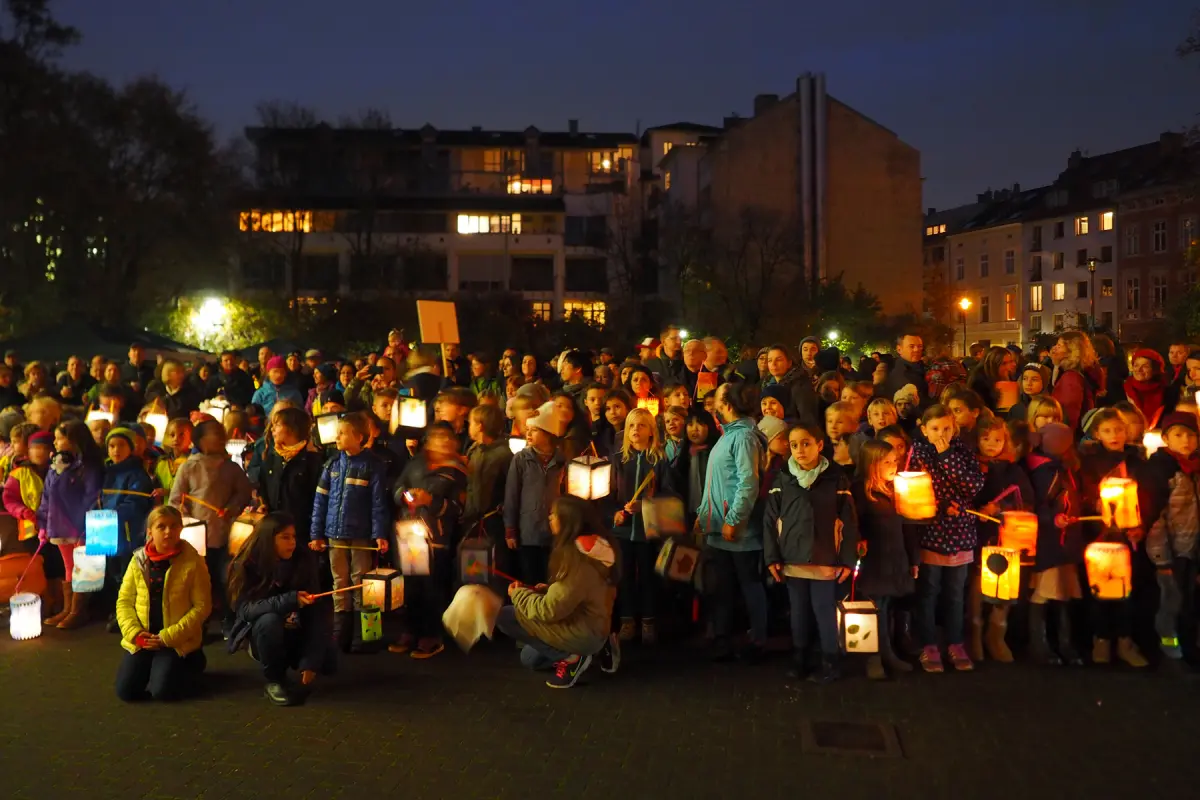 Kinder und Erwachsene mit leuchtenden Laternen bei Nacht auf einem Platz vor Wohnhäusern