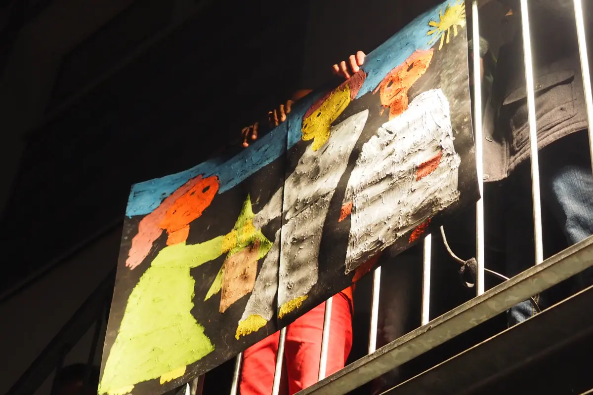 Bunt bemaltes Plakat mit kindlichen Figuren, über ein Metallgeländer gehängt, Hände und Beine dahinter sichtbar