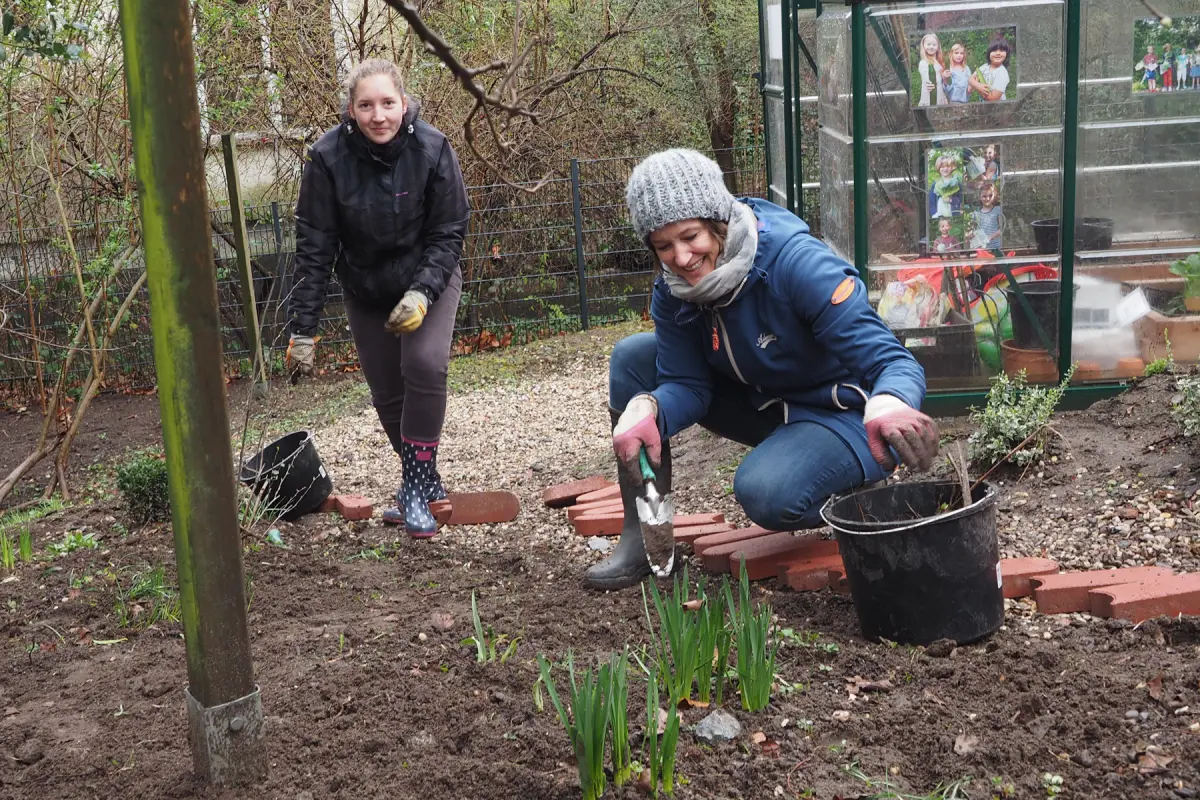 Zwei Personen beim Pflanzen im Garten; eine hockt mit Kelle neben Eimer, die andere steht mit Handschuhen und Gummistiefeln vor einem Gewächshaus.