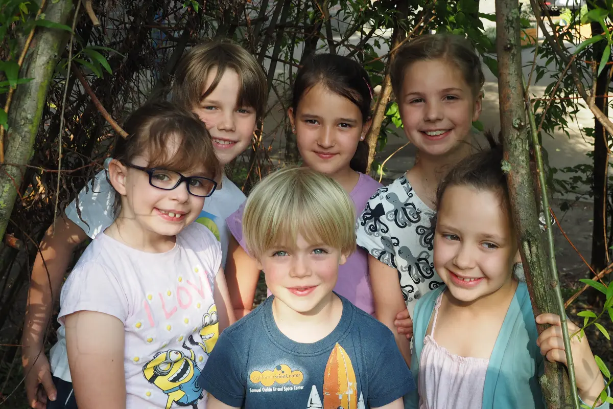 Sechs lächelnde Kinder dicht beieinander zwischen Ästen und Blättern im Freien.