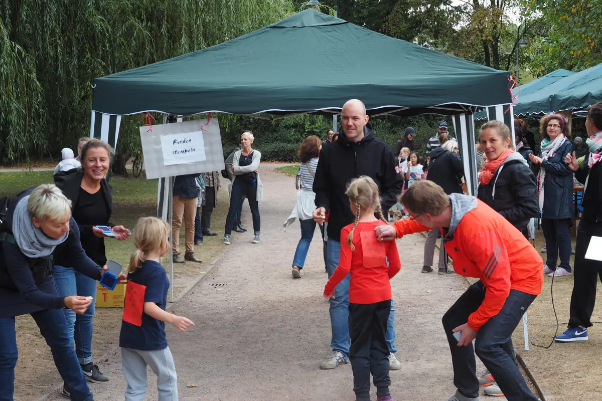 Kinder und Erwachsene unter grünem Pavillon, Mädchen bekommt Stempel auf den Rücken