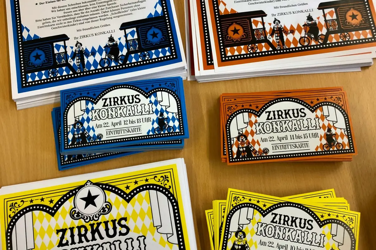 Gestapelte bunte Zirkus‑Flyer mit Schriftzug "ZIRKUS KONKALLI" in Blau, Orange und Gelb auf Holzuntergrund
